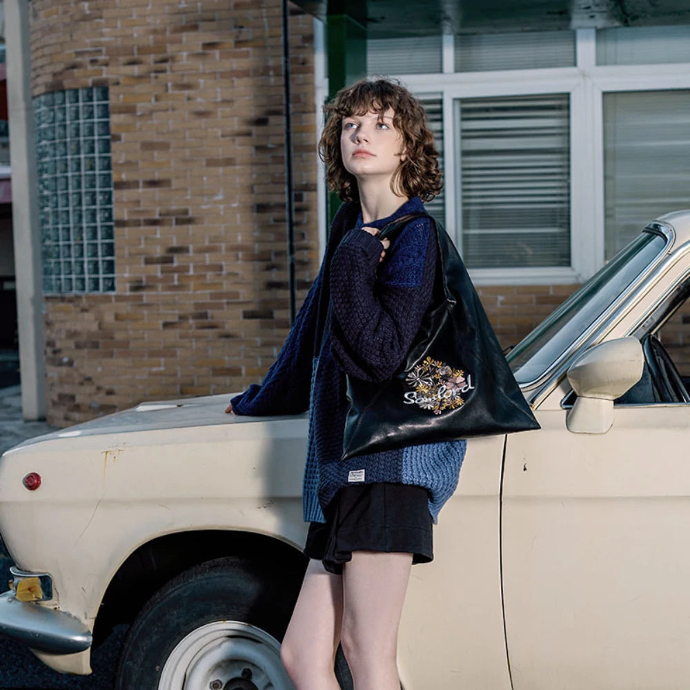 The model poses against an aged brick building, dressed in a cozy blue knit sweater, showcasing the Somlord Artistic Embroidered Triangle Soft Leather Tote Bag with Floral embroidery design. The soft leather and intricate stitching add a touch of autumn warmth.