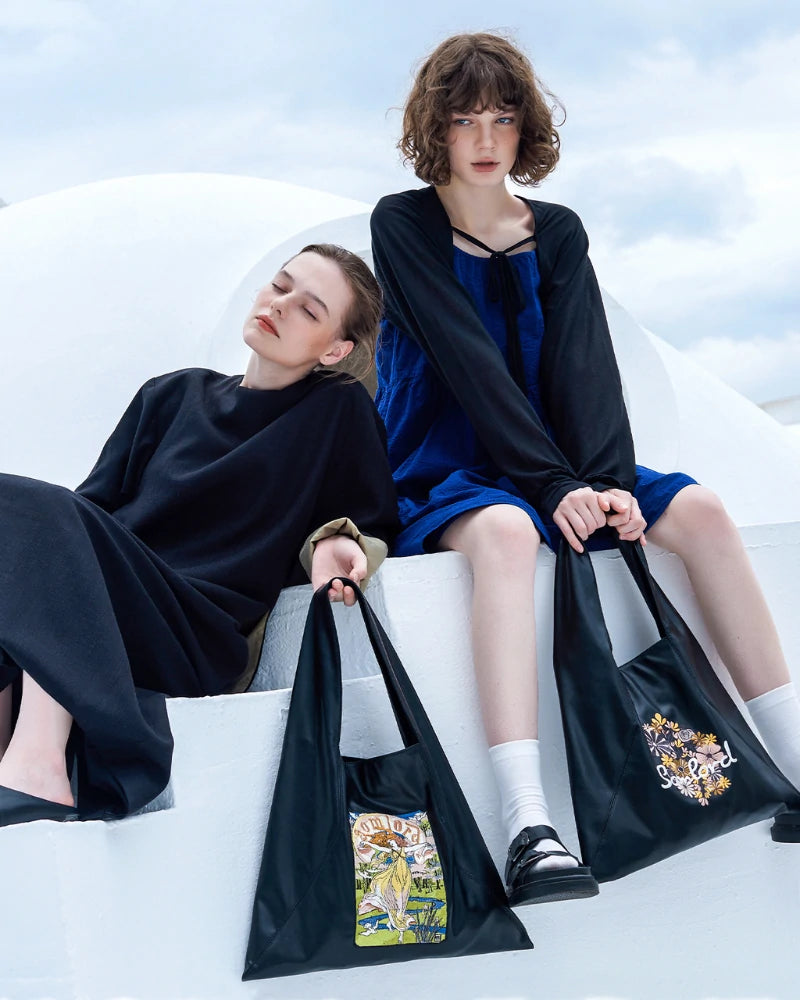 Two models sit side-by-side on white architecture steps, each carrying a Somlord Artistic Embroidered Triangle Soft Leather Tote Bag — one featuring the Goddess design and the other the Floral design. A perfect pairing of art and fashion.