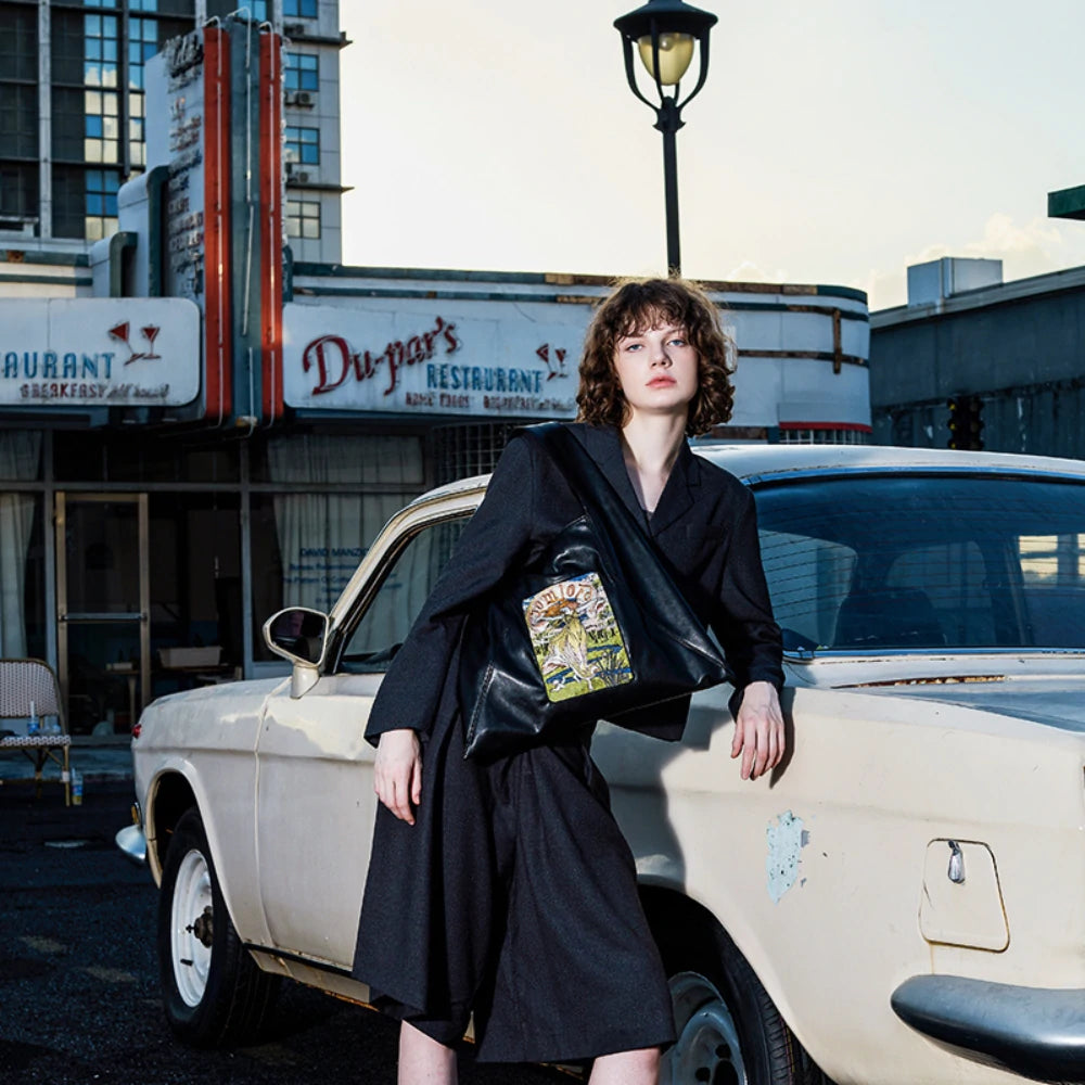 A model leans casually against a vintage car at sunset, wearing an elegant black suit and carrying the Somlord Artistic Embroidered Triangle Soft Leather Tote Bag with the Goddess embroidered design. The urban vintage setting enhances the sophisticated appeal of the bag.