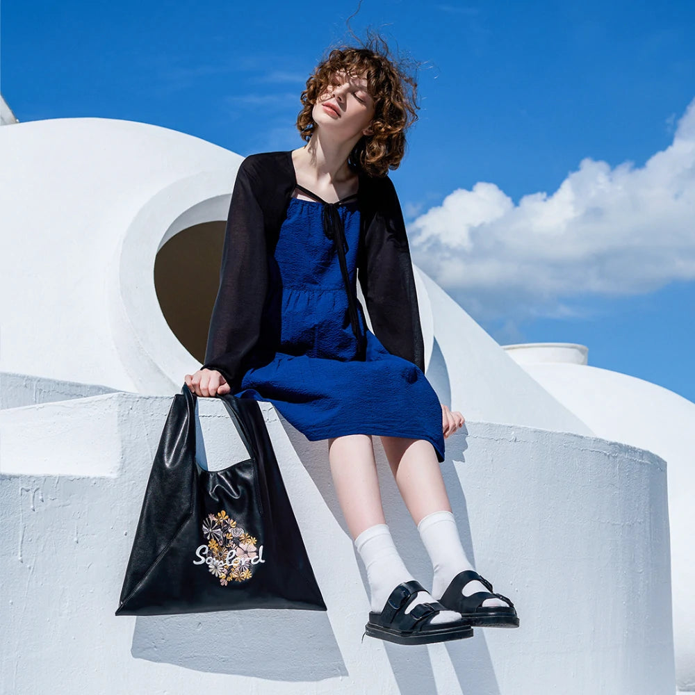 Under a bright blue sky, the model sits on a white modern architectural structure with the Somlord Artistic Embroidered Triangle Soft Leather Tote Bag featuring Floral embroidery design. The vibrant outdoor scene complements the bag’s artistic aesthetic.