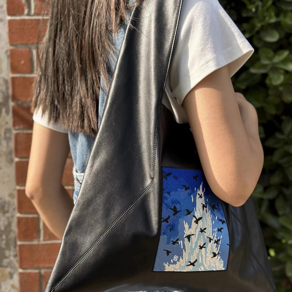 Sunlight glistens on the Somlord Artistic Embroidered Triangle Soft Leather Tote Bag slung over the model's shoulder, highlighting the Ocean Whisper embroidery design. The serene blue embroidery evokes a calm, ocean-inspired vibe.