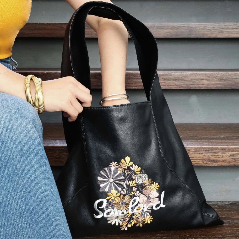 A close-up shot of the Somlord Artistic Embroidered Triangle Soft Leather Tote Bag resting on wooden stairs shows the Floral embroidery design in intricate detail. The model’s hand gently holds the tote, highlighting its artistic craftsmanship.